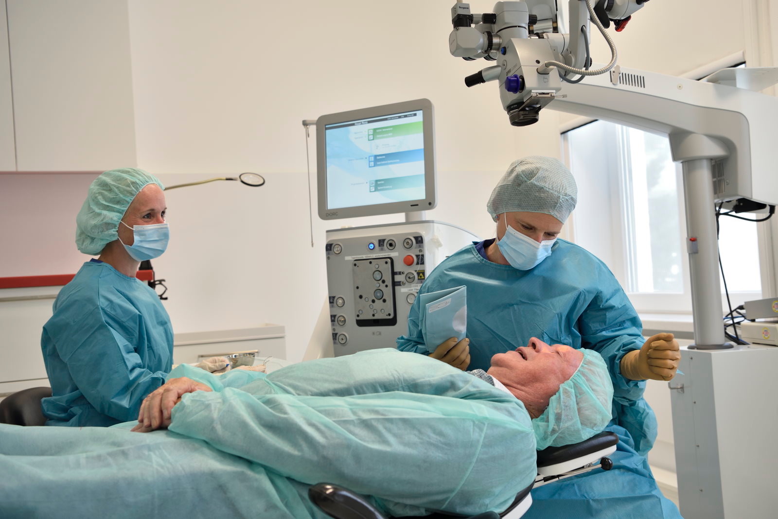 Zwei Mediziner in OP-Kleidung und mit Mundschutz bereiten einen Patienten in einem modernen Operationssaal auf einen Eingriff vor, wobei im Hintergrund medizinische Geräte und ein Monitor zu sehen sind.